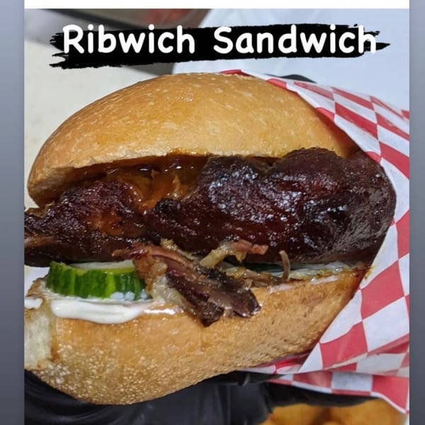 A Ribwich Sandwich with dark meat, pickles, and white sauce on a bun, wrapped in red and white checkered paper.