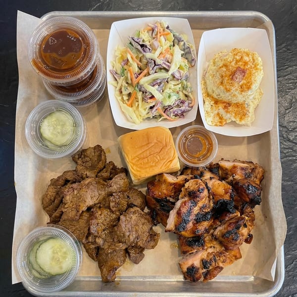 A metal tray with paper lining holds a meal of grilled chicken, beef, coleslaw, potato salad, pickles, sauces, and a bun.