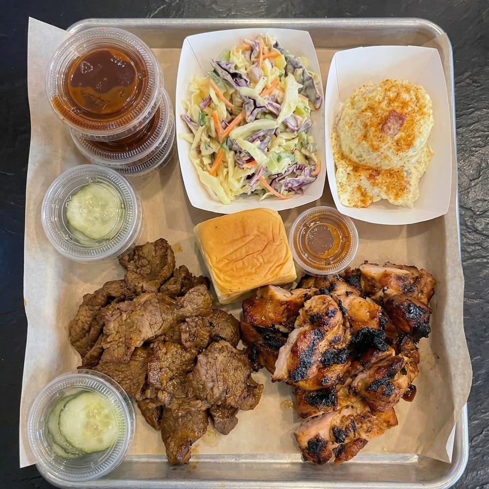 A metal tray with paper lining holds a meal of grilled chicken, beef, coleslaw, potato salad, pickles, sauces, and a bun.