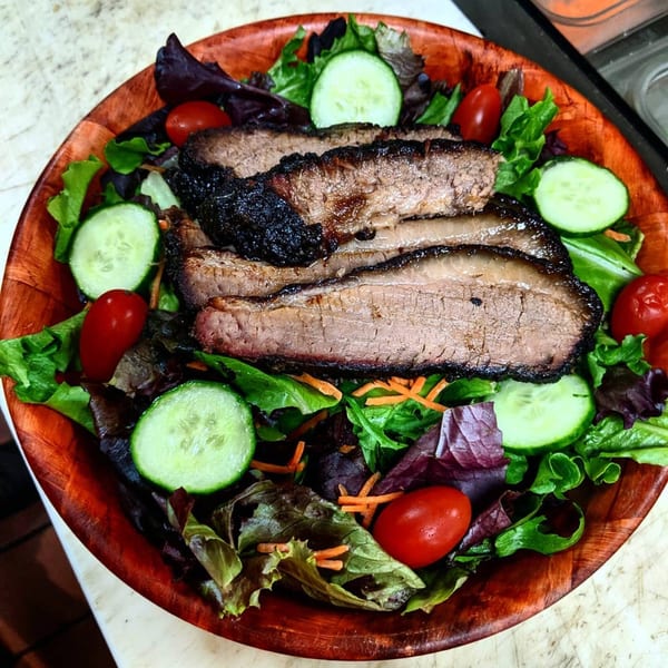 A wooden bowl filled with a fresh salad topped with sliced brisket, cucumbers, and cherry tomatoes.