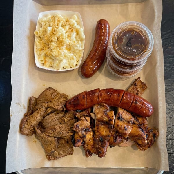 A BBQ platter with sliced brisket, grilled chicken, sausage, mac and cheese, and dipping sauces.