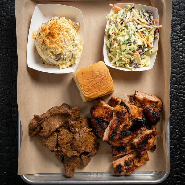 A tray with grilled chicken, sliced beef, potato salad, coleslaw, and a dinner roll.